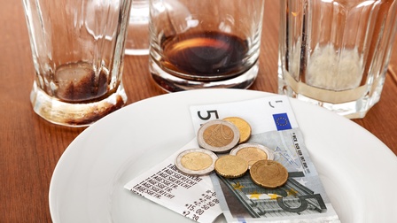 Nahaufnahme eines runden, kleinen Tellers, auf dem eine Rechnung, ein Fünf-Euroschein sowie ein paar Münzen liegen. Daneben stehen vier Gläser