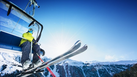 Person in Wintersportbekleidung sitzt mit Skiern auf einem Sessellift während sich im Hintergrund ein Bergpanorama erstreckt