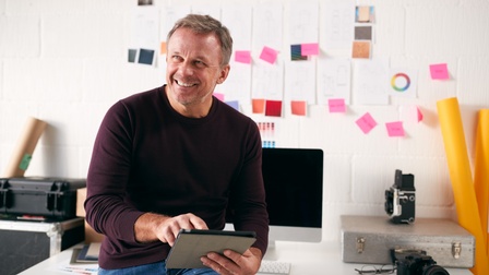 Lächelnde Person sitzt auf Schreibtisch und bedient Tablet, im Hintergrund Fotografieutensilien und Post-its an der Wand