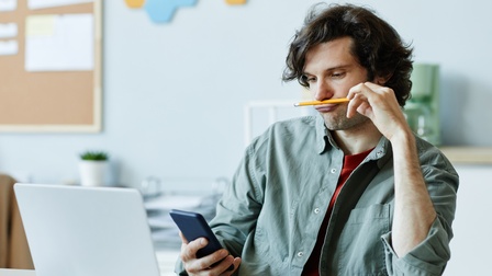 Eine Person sitzt an einem Tisch. Vor ihr ist ein aufgeklappter Laptop. Die Person hält ein Smartphone, auf das sie blickt. Mit der linken Hand hält sie einen Bleistift zwischen Nase und Oberlippe