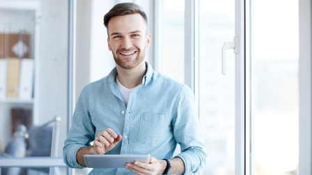 Portrait einer Person mit Bart und blauem Hemd, die freudig ein Tablet in einem hellen Büro in Händen hält
