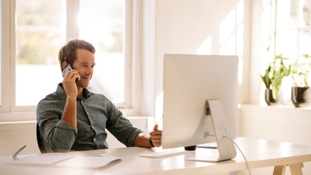 Person sitzt am Computer und telefoniert mit Smartphone