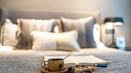Auf der Decke eines gemachten Bettes sind eine Tasse, ein Operngucker und ein aufgeklapptes Buch. Im Hintergrund sind mehrere Pölster sowie ein Nachtkästchen mit einer Stehlampe