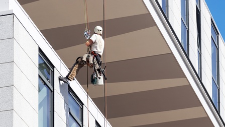 Eine Person mit Helm seilt sich von einem Gebäude hab und hat rechts neben sich Putzutensilien hängen