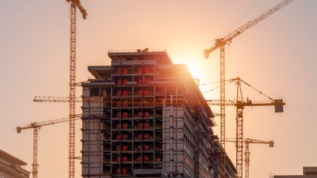 Ein mehrgeschossiges Gebäude befindet sich im Bau. Die Tragkonstruktion und mehrere Geschosse sind freigelegt. Rund um das Gebäude stehen mehrere Baukräne. Die Sonne befindet sich hinter dem oberen Gebäudebereich