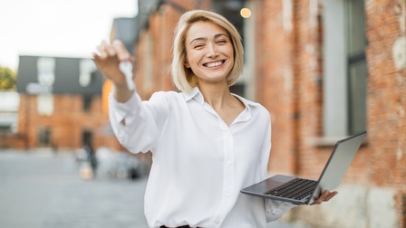 Porträt einer Person mit weißer Bluse, kinnlangen, hellen Haaren und aufgeklappten Laptop in der linken Hand, die neben einem Haus steht. Mit der rechten Hand hält sie einen Schlüssel in die Kamera und lächelt