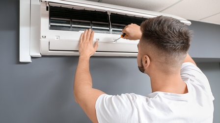 Rückenansicht einer Person, die die Abdeckung einer Klimaanlage geöffnet hat. Die Person hält einen Schraubenzieher auf die Klimaanlage.