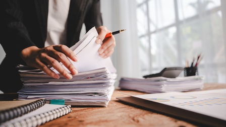 Eine Person im Ausschnitt mit Stift zwischen den Fingern blättert durch einen Stapel Dokumente, die auf einem Tisch liegen.