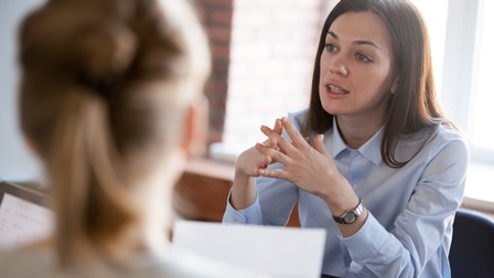 Person mit langen dunklen Haaren, hellblauer Bluse und silberner Uhr spricht mit anderen Personen in der Unschärfe