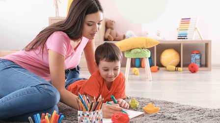 Ein Kind liegt in Bauchlage auf einem grauen Teppich. Links neben dem Kind sitzt eine erwachsene Person. Beide halten einen Stift in der rechten Hand und malen in ein offenes Heft. Vor ihnen stehen weitere Boxen mit Stiften