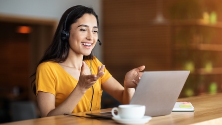 Person mit Headset und gelbem T-Shirt sitzt freudig vor einem Laptop und hält eine Konferenz, daneben steht eine Tasse sowie liegt ein Notizbuch auf dem Holztisch in einem Café