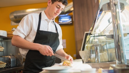 Seitliche Aufnahme einer Person mit kurzen, dunklen Haaren und Schürze, die an einem Tresen neben einer Kühltheke steht und mit einem Brotmesser ein Brötchen schneidet