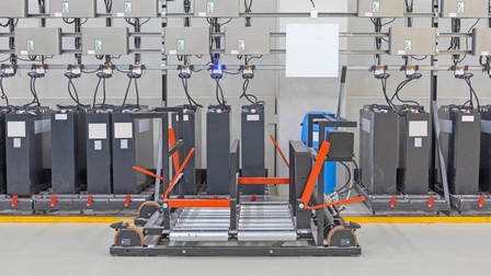 Reihe grauer Industriebatterien verbunden mit Kabeln zu grauen Boxen, im Vordergrund Hebegerät