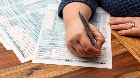 Nahaufnahme von Händen einer Person, die linke Hand  liegt auf einem Holztisch, in der rechten hält sie einen Kugelschreiber. Mit diesem füllt sie ein Formular, das neben anderen Dokumenten auf dem Tisch platziert ist