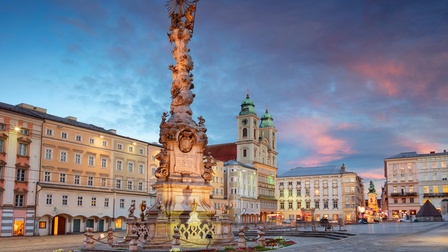 Die Pestsäule in Linz auf dem Hauptplatz bei Abenddämmerung. Der Platz wird von verschiedenen Altbauten und Kirchen gesäumt