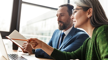 Zwei Personen sitzen nebeneinander an einem Tisch. Vor ihnen ist ein Laptop. Die rechte Person mit Brille und langem, offenem Haar deutet mit einem Bleistift darauf. Die linke Person mit Bart hält ein Notizbuch