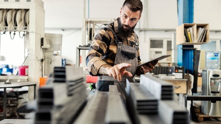 Person in kariertem Hemd und Arbeitskleidung steht in einer Werkstatt und kontrolliert Betriebsmittel während ein Tablet in der Hand gehalten wird