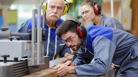Drei Personen mit Hörschutz an Sägemaschine in Arbeitsoveralls, eine Person schneidet Holzstück zu