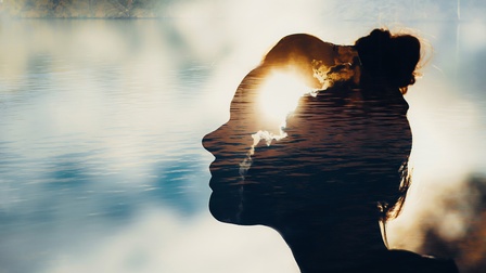 Doppelbelichtungsfoto, das die Silhouette eines menschlichen Kopfes im Profil zeigt. Im Hintergrund ist eine harmonische Landschaft aus einem See und Bäumen. Der Kopf ist mit der Spiegelung des Himmels und der Wolken auf dem Wasser gefüllt