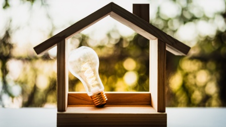 Miniaturholzhaus steht bei einem Fensterbrett und darin leuchtet eine  angelehnte Glühbirne, im Hintergrund zeigt sich ein grün, gelbes Bokeh