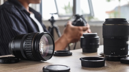 Eine Person sitzt an einem Tisch und hält einen Fotoapparat. Auf dem Tisch sind verschiedene Kameraobjektive und -linsen