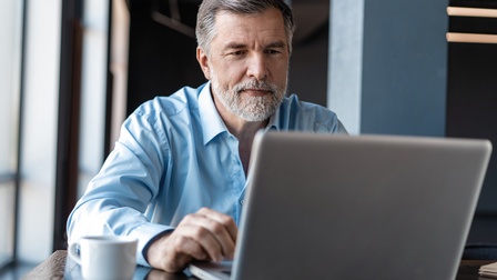 Person mit kurzen weiß-grauen Haaren und Bart mit blauem Hemd sitzt freudig vor einem Laptop in einem modernen Büroraum