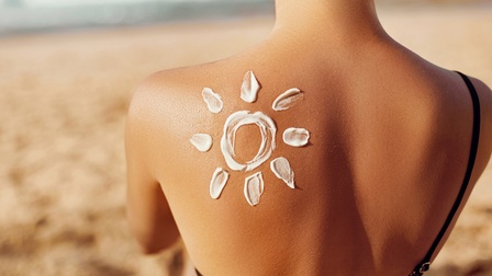 Eine Sonnencreme ist in Form einer Sonne aufgetragen und zeigt sich auf der Schulter einer Person in Bikini, die bei einem Strand sitzt