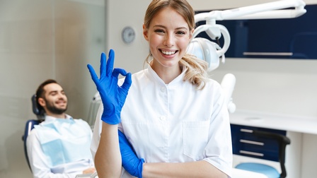 Eine Person in weißem Arbeitskittel steht lachend in einer Zahnarztpraxis und macht mit einer Hand ein OK-Zeichen. Im Hintergrund liegt eine weitere Person auf einem Zahnarztstuhl.