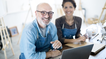 Zwei lächelnde Personen mit Schürzen sitzen vor Laptop, ringsum Malerutensilien