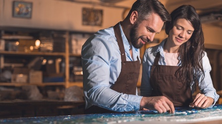 Zwei Personen mit sanften Lächeln und Arbeitsschürzen tragend legen Mosaiksteine