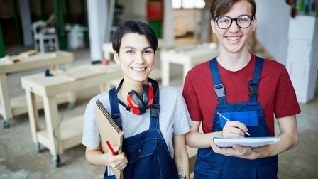 Zwei lächelnde Personen in blauen Arbeitslatzhosen mit Stiften und Notizblöcken in Tischlerwerkstatt stehend