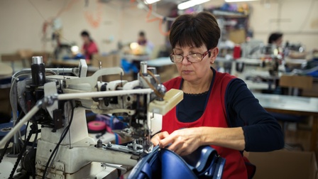 Frontalaufnahme einer hinter einer weißen Nähmaschine sitzenden Person mit Brille, roter Schürze und dunklem Oberteil. Sie hält mit beiden Händen ein blaues, gummiartiges Objekt an die Nähmaschine. Im Hintergrund sitzen weitere Personen