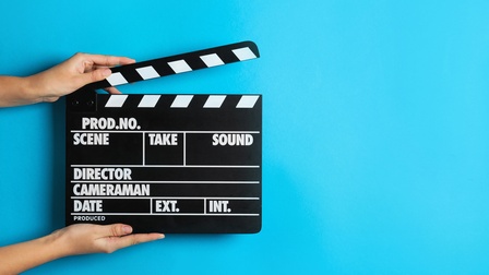 Filmklappe auf blauem Hintergrund von zwei Hände gehalten: Schwarzweiße Platte mit den Aufschriften Scene, Take, Sound und weiteres mit beweglichem Klappteil an der oberen Kante