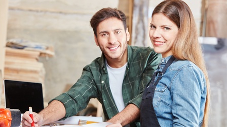 Zwei Personen stehen nebeneinander an einem Holztisch und blicken in die Kamera. Die rechte Person trägt eine Schürze. Die linke Person zeichnet mit einem Bleistift etwas auf ein großes Blatt Papier. Daneben liegt ein Maßband