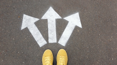 Gelbe Schuhe stehen auf einem Asphalt, davor sind drei weiße Pfeile, die in unterschiedliche Richtungen zeigen am Boden markiert