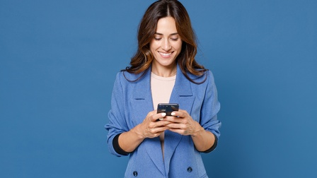 Person mit langen braunen Haaren und blauem Blazer blickt lächelnd auf Smartphone, das sie in Händen hält, blauer Hintergrund