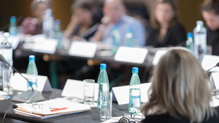 Um einen Tisch sitzen mehrere Personen. Vor jeder Person steht ein Kärtchen, ein Glas Wasser und eine Plastikflasche. Vor manchen Personen sind Notizblöcke und ein Mikrofon