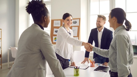Vier Personen in formaler Kleidung stehen in einem Raum um einen Tisch. Zwei Personen stehen sich gegenüber und schütteln sich die Hände