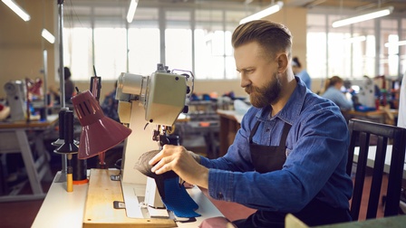 Seitliche Aufnahme einer Person mit Bart, kurzen Haaren und Schürze, die an einem Tisch in einer Werkstatt sitzt und eine Nähmaschine bedient. Im Hintergrund sitzen weitere Personen an Nähmaschinen