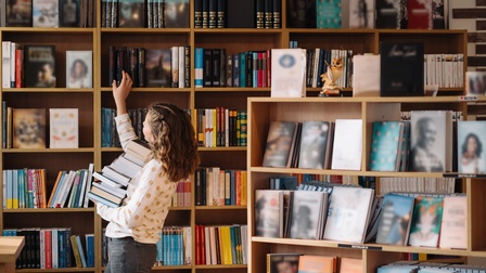 Eine Person hat am linken Arm einen hohen Bücherstapel. Sie steht seitlich vor einem langen Regal mit vielen Fächern. In jedem Fach sind viele Bücher. Die Person hat die rechte Hand auf einem Buchrücken. Im Vordergrund sind weitere Regale mit Büchern