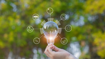 Glühbirne wird von einer Hand in die Höhe gehalten und diverse Icons zum Thema Energiesparen und Nachhaltigkeit strahlen darum, im Hintergrund zeigt sich ein florales Bokeh aus gelb und grünen Farben