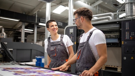 Zwei Personen in Arbeitslatzhosen stehen an Tisch mit Druckbögen und blicken einander an, im Hintergrund große Druckmaschine