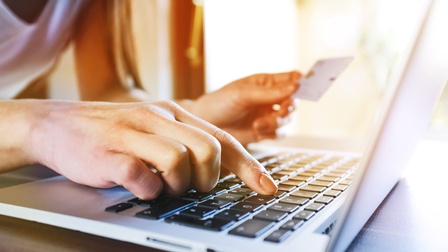 Detailaufnahme einer Person, die mit dem rechten Zeigefinger auf eine Buchstabentaste eines Laptops tippt während in der linken Hand eine Karte gehalten wird.