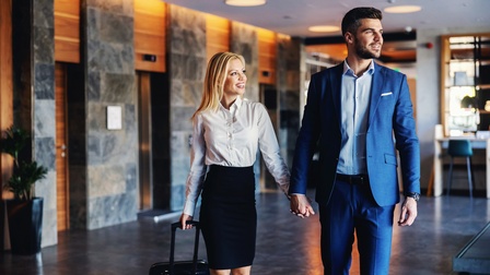 Zwei lächelnde Personen in Hotellobby Hand in Hand gehend, eine Person zieht Rollkoffer nach, im Hintergrund Lifte