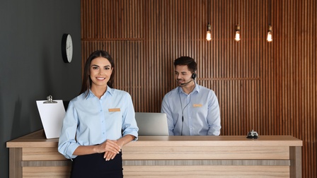 Porträt einer Person mit Namensschild, die sich an einer Hotelrezeption abstützt. Hinter der Rezeption steht eine Person mit Headset, die auf den Bildschirm eines Computers blickt