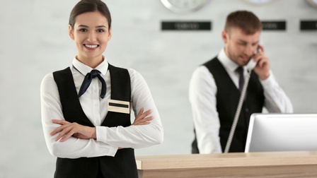 Person in Arbeitskleidung steht vor einem Empfang mit verschränkten Armen und freundlichen Blick, im Hintergrund steht eine weitere Person in Arbeitskleidung, die telefoniert und dabei auf einen Bildschirm-Monitor blickt