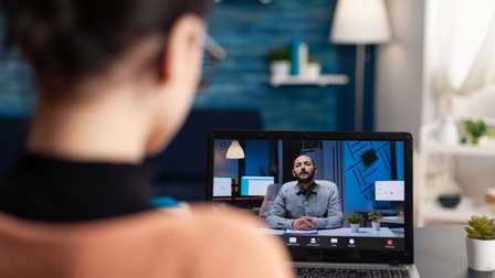 Person in Rückenansicht sitzt vor aufgeklapptem Laptop auf dessen Monitor weitere Person zu sehen ist