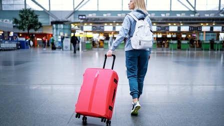 Rückenansicht einer Person, die gerade einen Flughafen betritt und ihren roten Koffer mit sich zieht. Am Rücken trägt sie einen weißen Rucksack und schaut auf die Seite.
