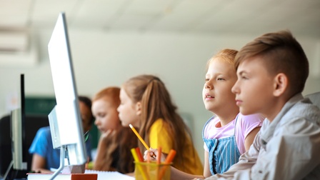 Seitliche Aufnahme von fünf Kindern in einer Schulklasse, die nebeneinander an Tischen sitzen und in Gruppen auf Bildschirme blicken, die vor ihnen stehen.