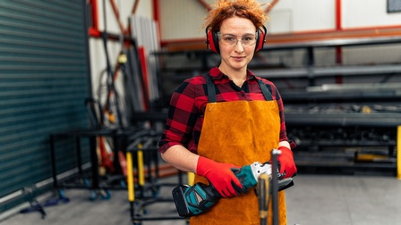 Porträt einer lächelnden Person mit Schutzbrille, Gehörschütz, Handschuhen, Schürze und geschlossenen Haaren, die in einer Werkstatt steht und ein Flexgerät hält